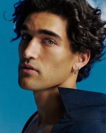 Portrait of a young adult male model with dark hair, light eyes, wearing silver earrings and a dark blue shirt against a light blue background.