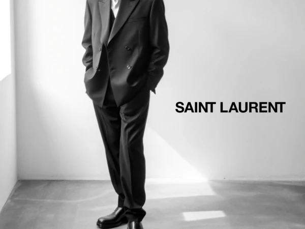 Headless model wearing an oversized black double-breasted suit and leather dress shoes standing beside a bold Saint Laurent logo.