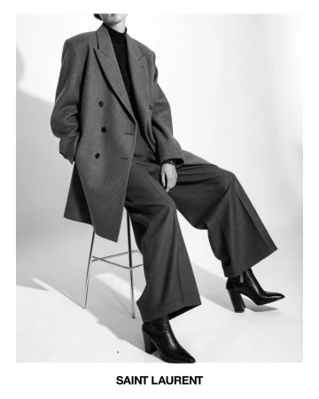 Model seated on a stool wearing an oversized grey double-breasted suit, black turtleneck, and heeled leather ankle boots in a studio.