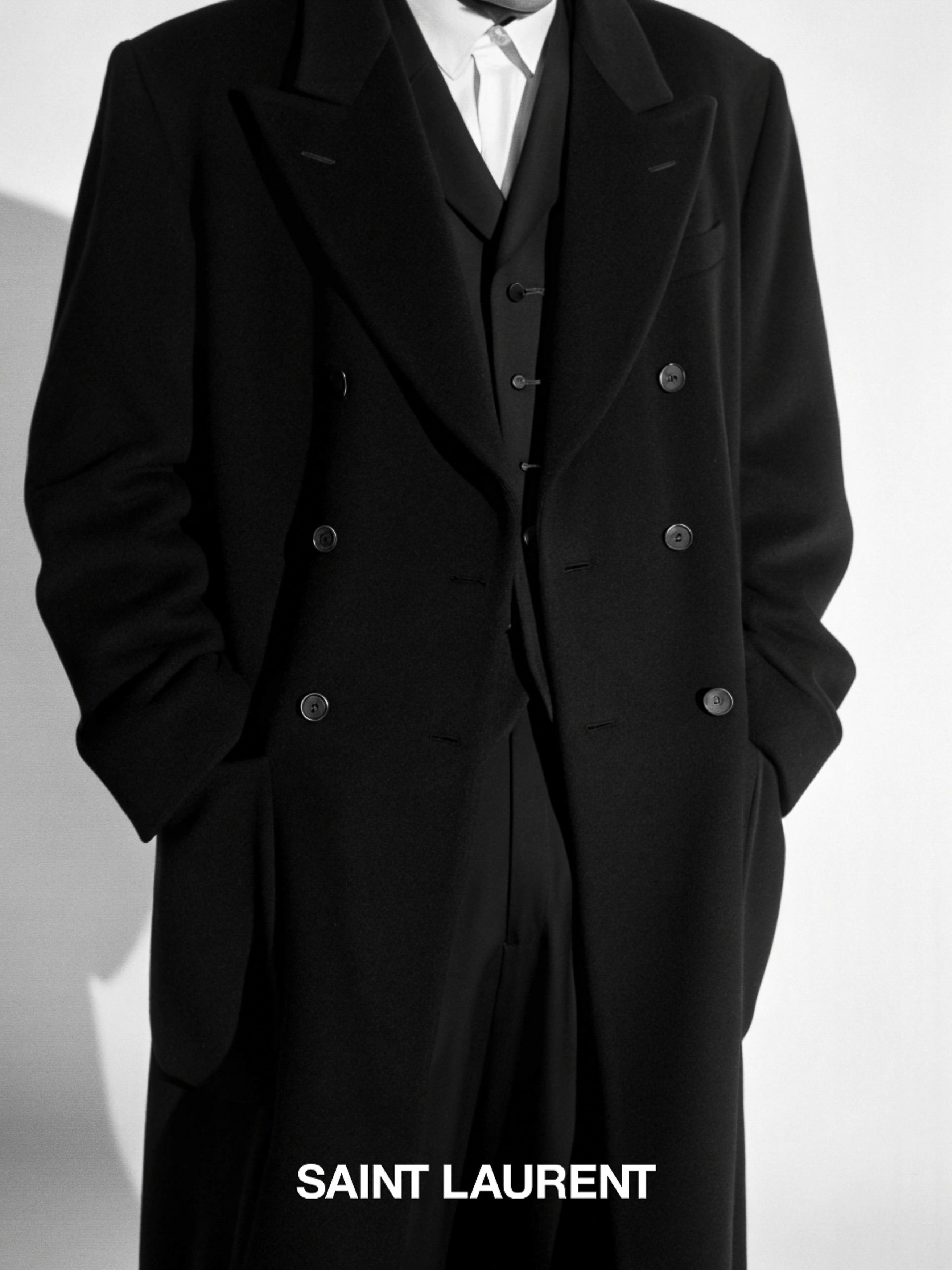Person in a black wool overcoat, vest, and trousers from Saint Laurent, standing in a neutral setting with natural light.