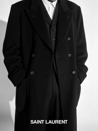 Person in a black wool overcoat, vest, and trousers from Saint Laurent, standing in a neutral setting with natural light.