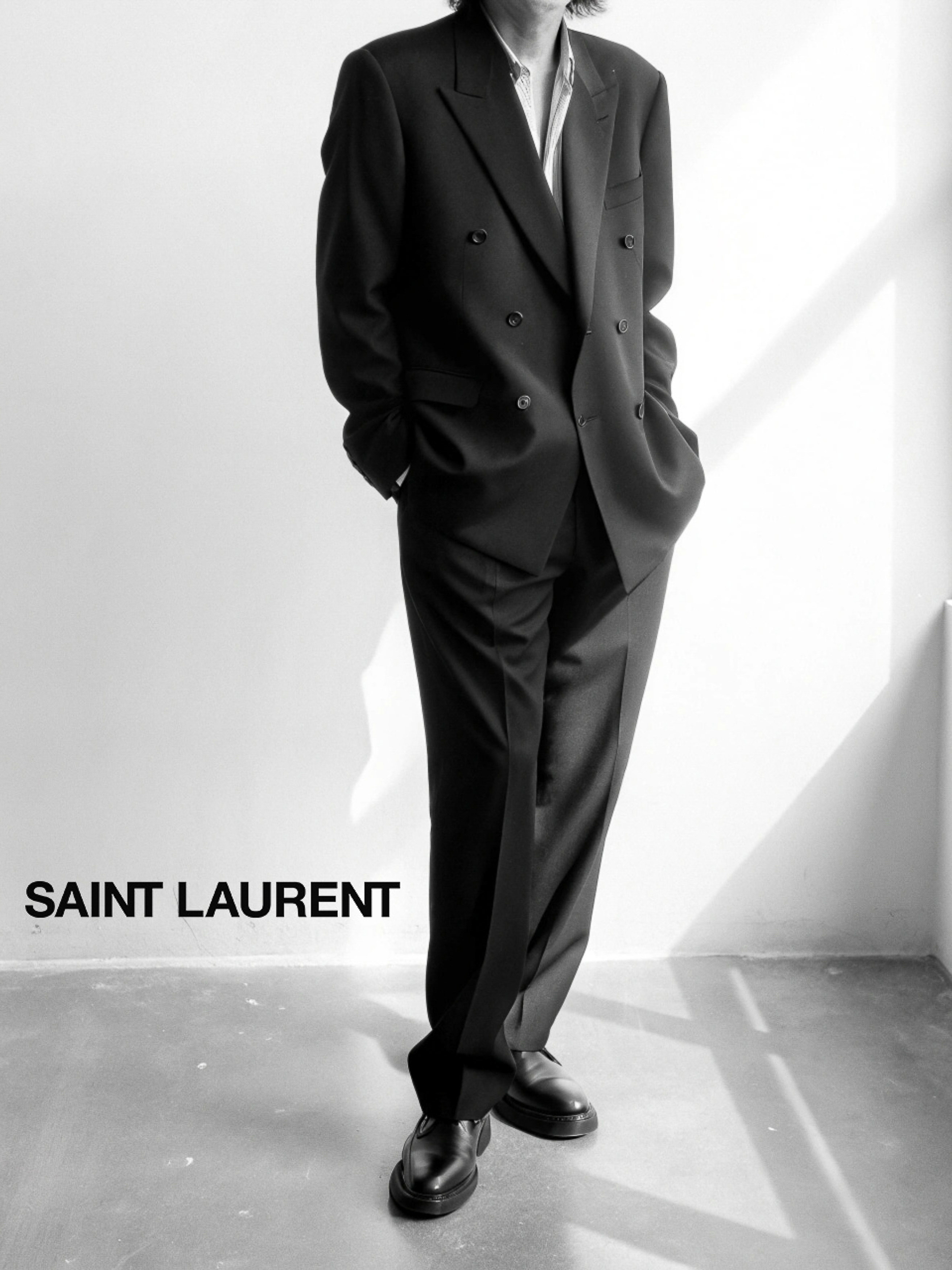 Monochromatic image featuring an adult male wearing a tailored black suit with a double-breasted jacket and dress shoes, standing indoors.