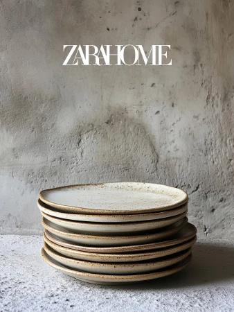 Stack of round speckled stoneware plates against textured grey wall, product shot for Zara Home, minimalist tableware.
