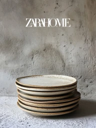 A stack of speckled cream ceramic dinner plates resting against a rustic, textured concrete wall with the Zara Home logo.