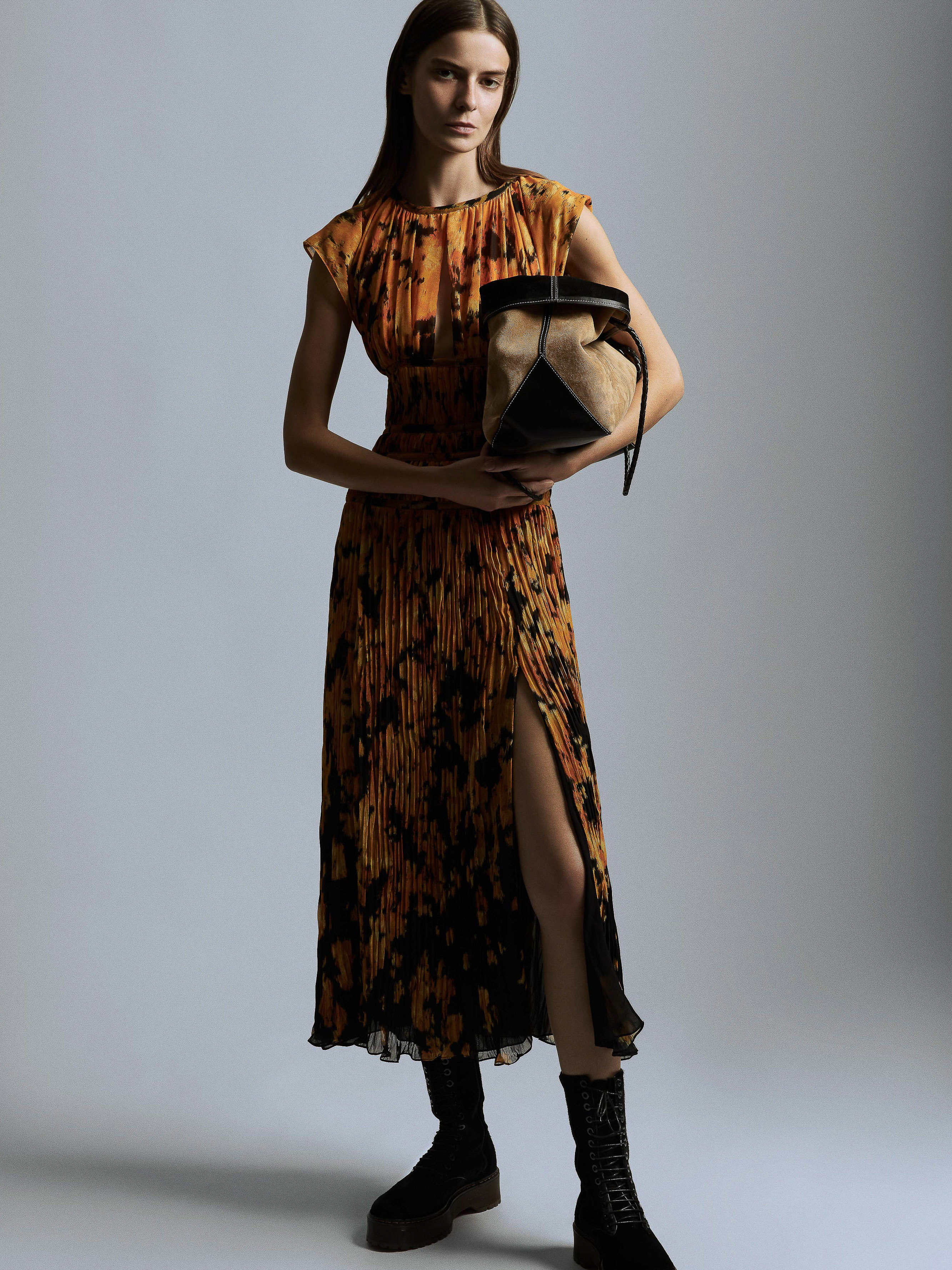 A fashionable adult woman poses in a black and orange dress, holding a beige and black purse, in a studio with a grey backdrop.