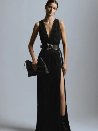 Fashion model in a black sleeveless gown with a thigh-high slit, accessorized with a wide belt and clutch bag, poses in a studio.