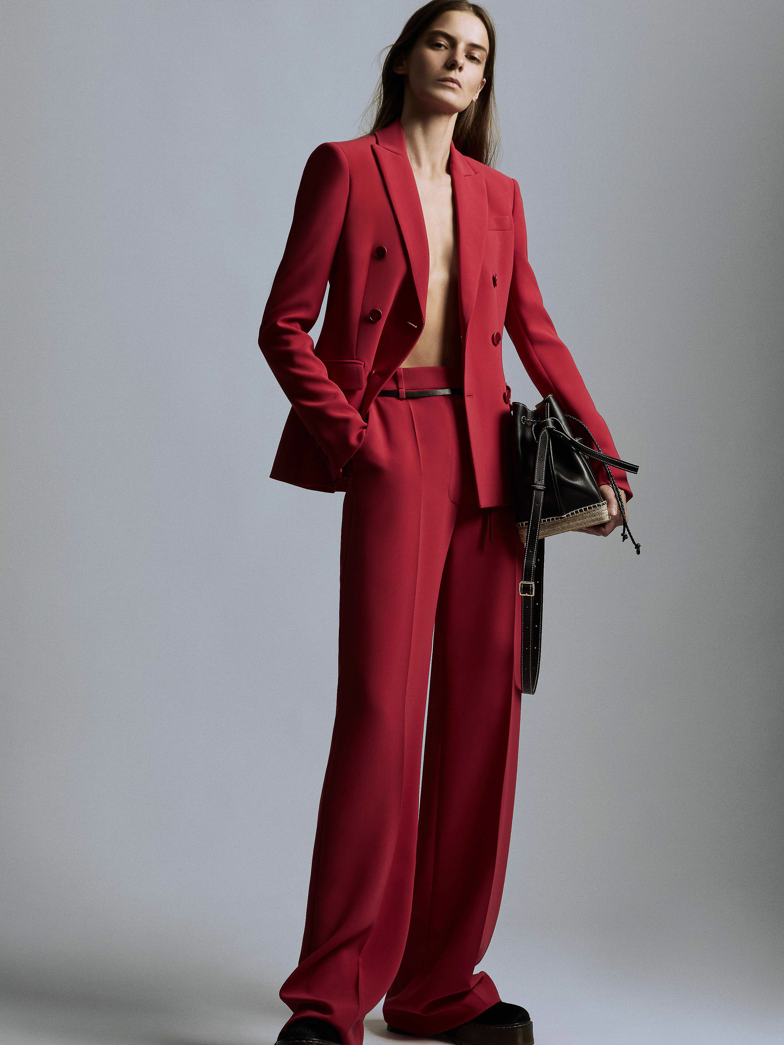 Fashion editorial shot of a model posing in a red double-breasted blazer, wide-leg trousers, and platform shoes against a grey backdrop.