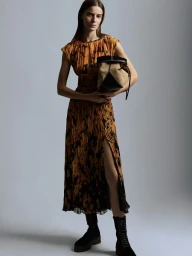 Model in a patterned pleated dress holding a bucket bag and wearing dark boots against a grey background