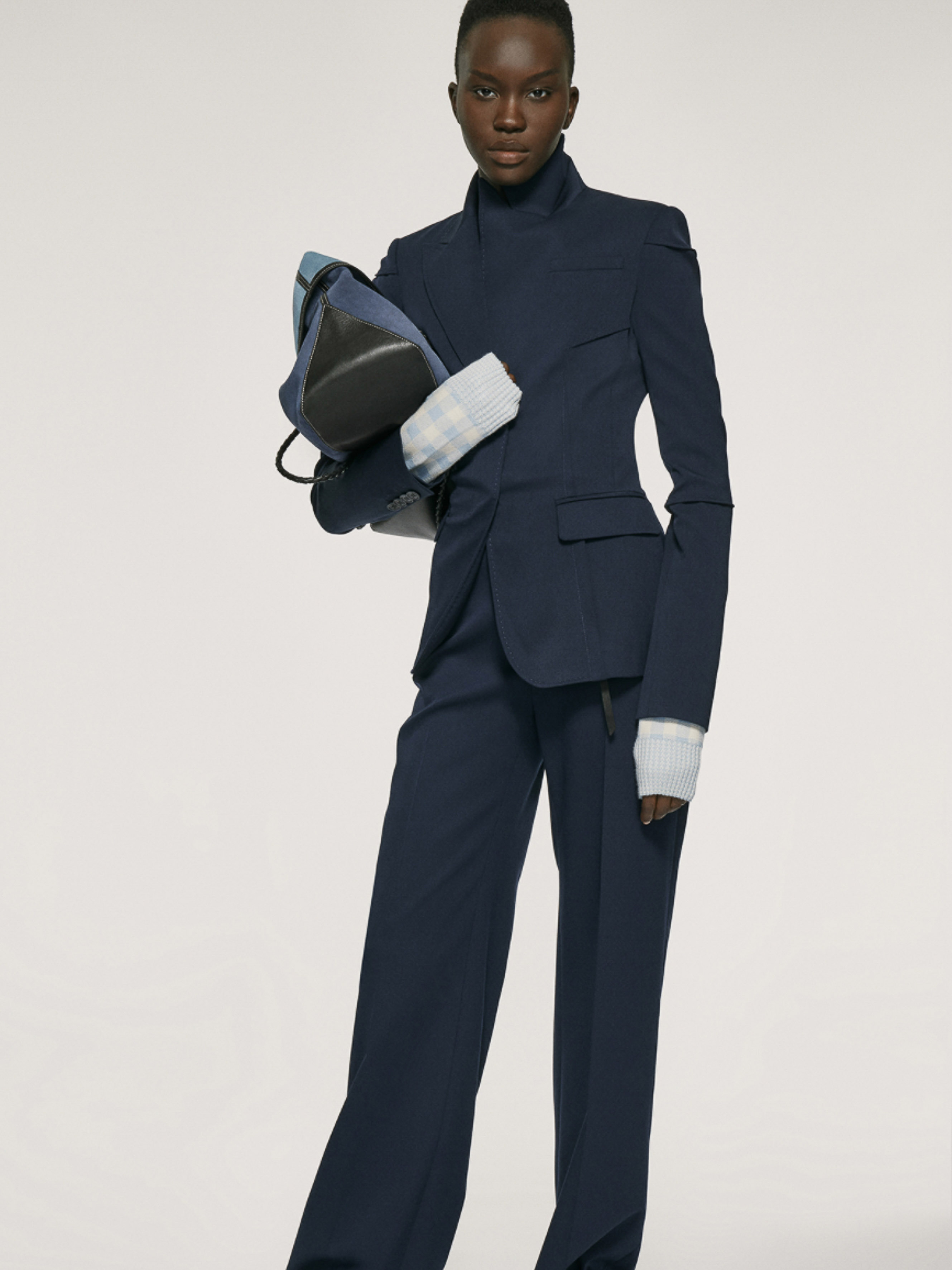 Dark-skinned female model in a navy blue pantsuit, holding a backpack in a studio setting, presenting a modern, professional fashion aesthetic.