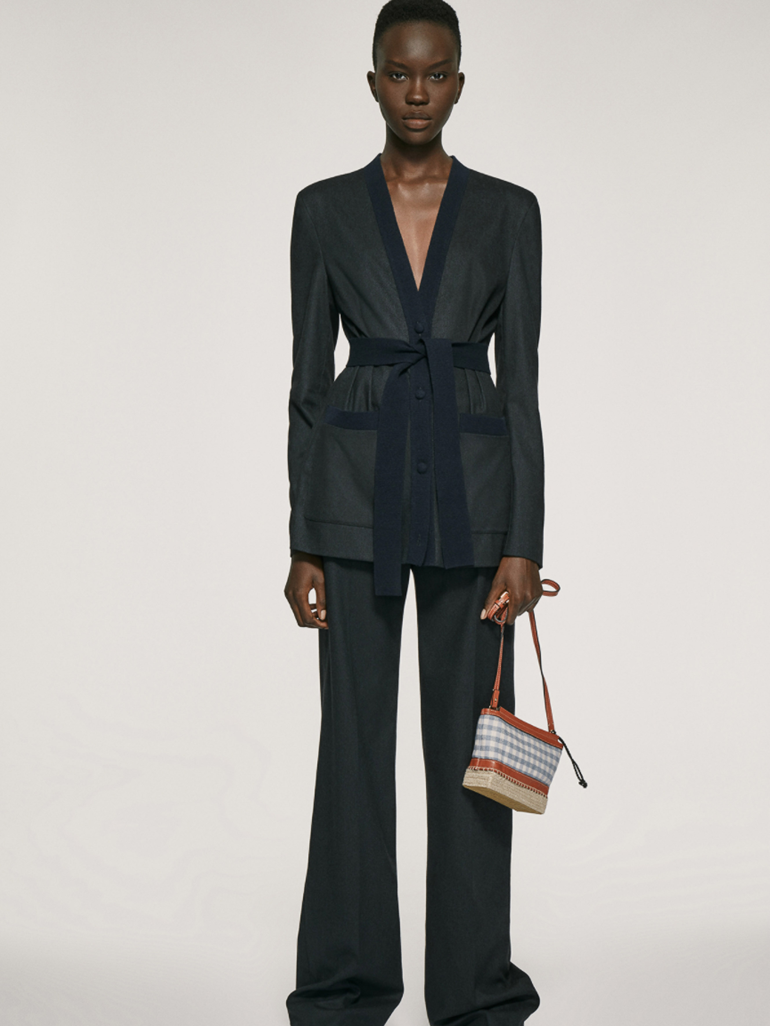 Model wearing a dark tailored belted blazer suit and wide-leg trousers while holding a small patterned handbag against a studio backdrop.