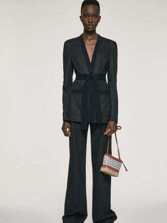 Model wearing a dark tailored belted blazer suit and wide-leg trousers while holding a small patterned handbag against a studio backdrop.