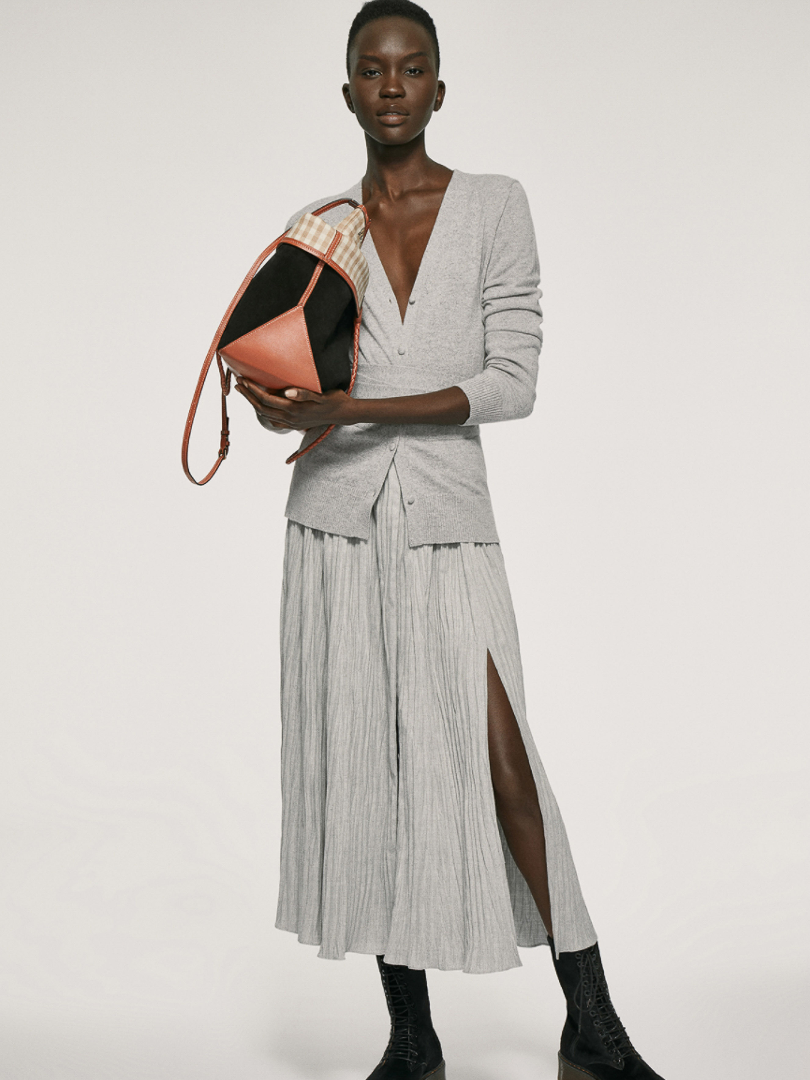 Fashion editorial shot of a Black woman wearing a grey cardigan and pleated skirt, holding a geometric color-blocked handbag.