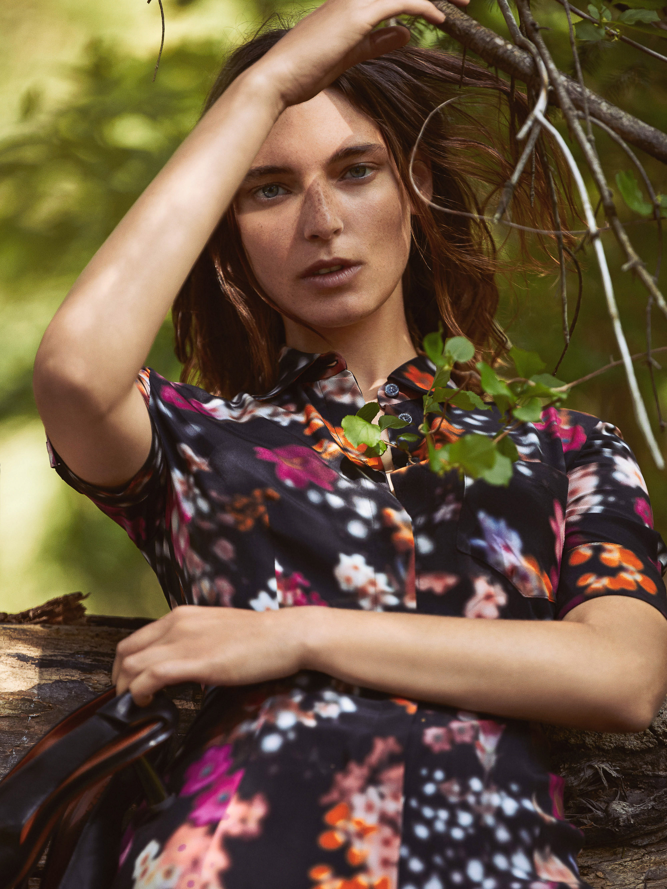 Woman in a floral dress poses in a sunlit forest scene. The photograph captures a blend of fashion and nature with soft, diffused lighting.