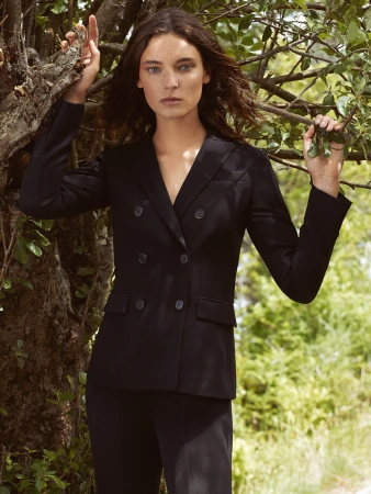 Brunette woman in a black double-breasted blazer and pants posing outdoors surrounded by lush green foliage and tree branches.