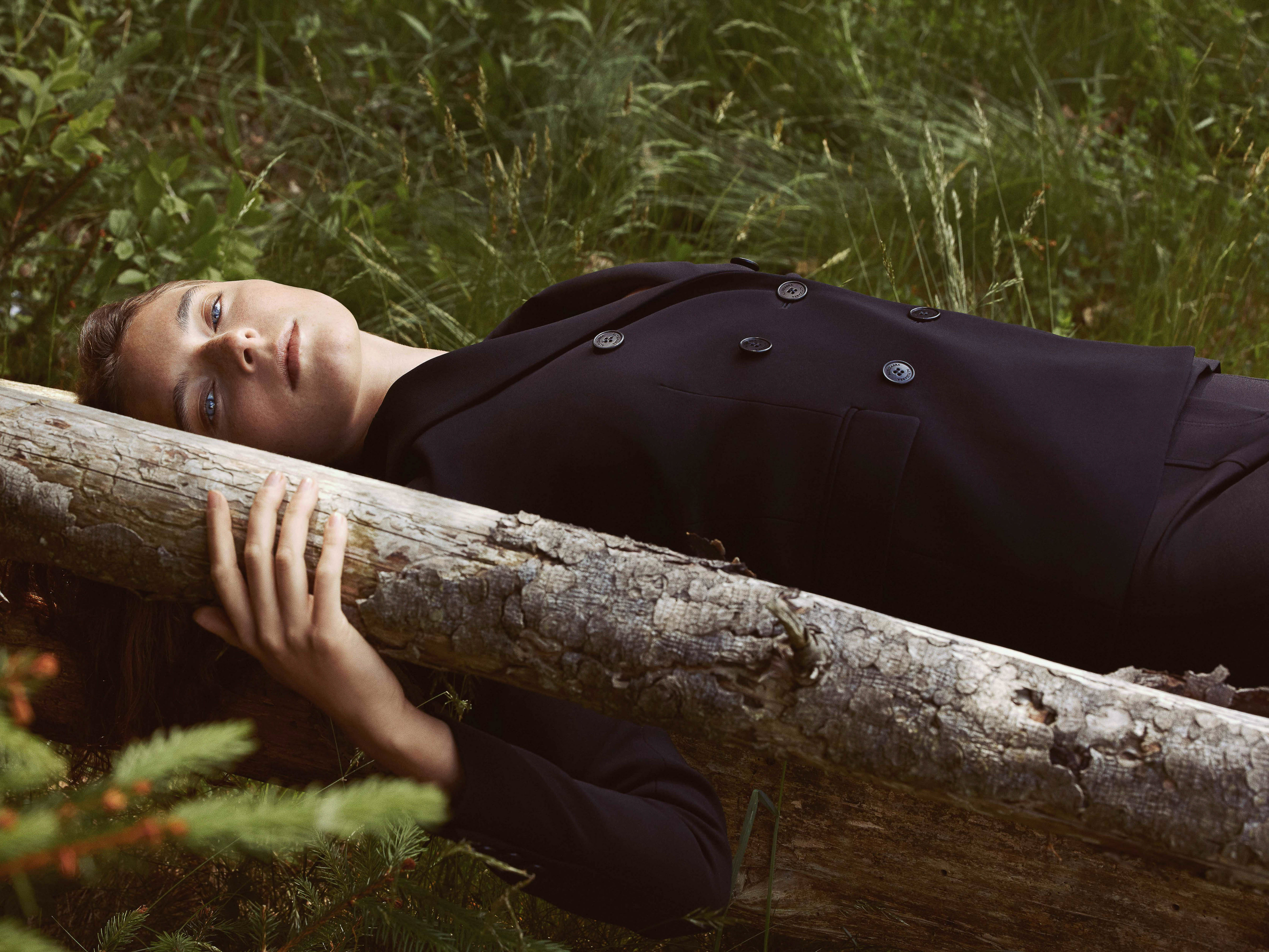 A woman in a dark suit lies on a log in a grassy forest, eyes open and gazing upward, in a fashion or beauty editorial style.