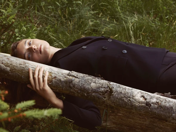 A woman in a dark suit lies on a log in a grassy forest, eyes open and gazing upward, in a fashion or beauty editorial style.