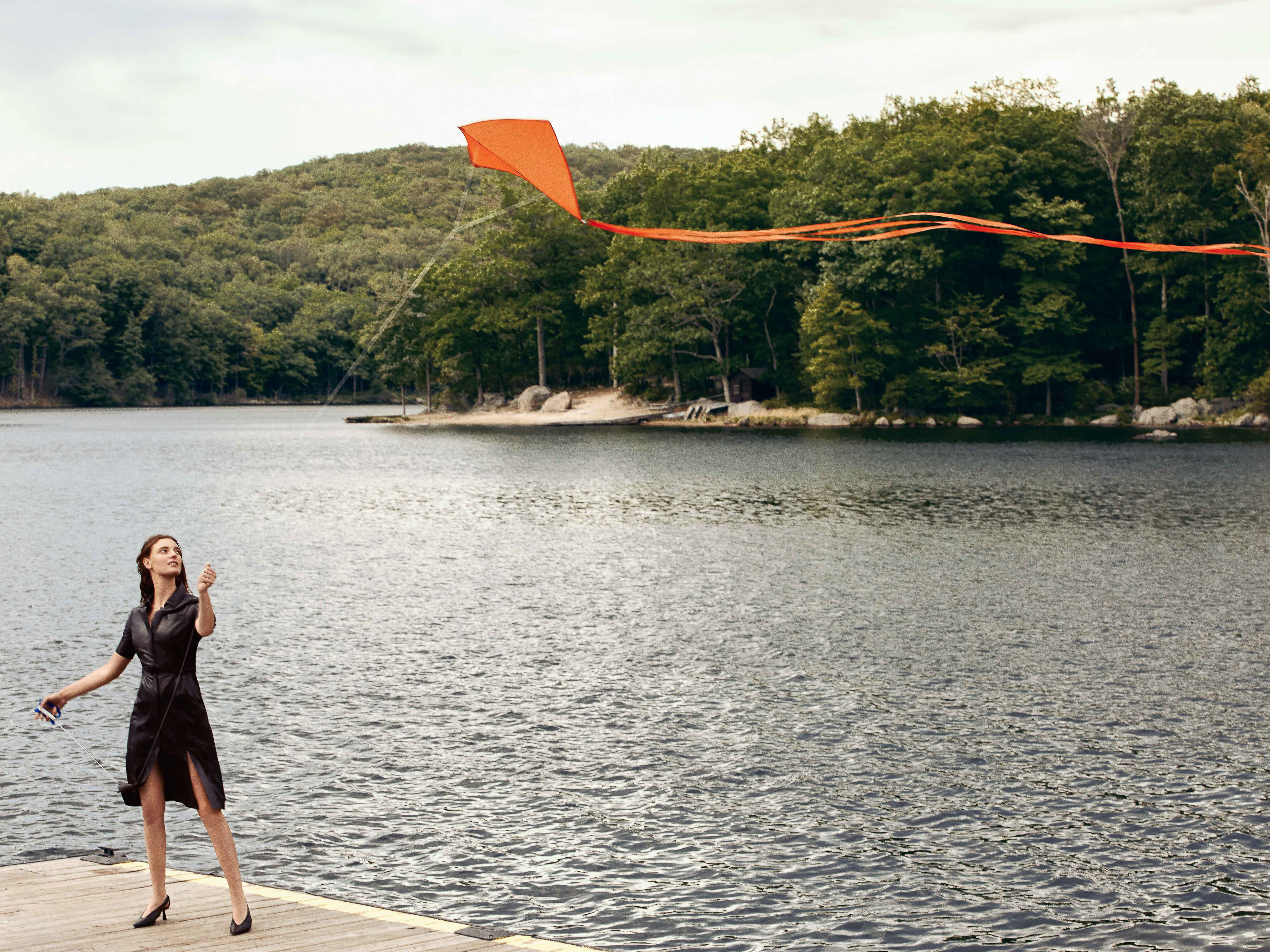 Adult woman in dress flies a kite on a wooden dock by a lake with a wooded shoreline on a cloudy day. Fashion shoot.