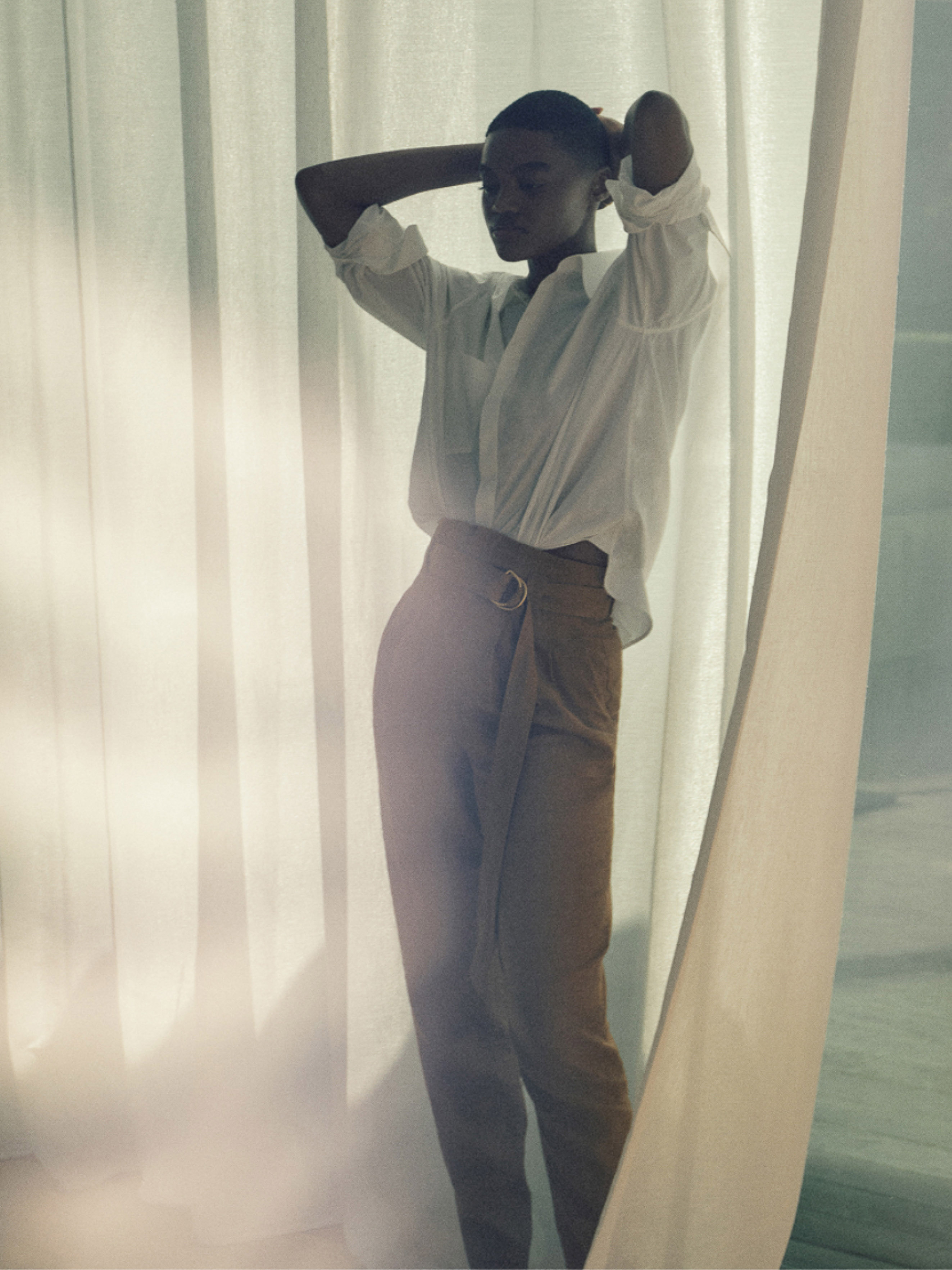 Fashion shot of a woman wearing a white blouse and khaki pants, posing with her hands behind her head against a curtain backdrop.