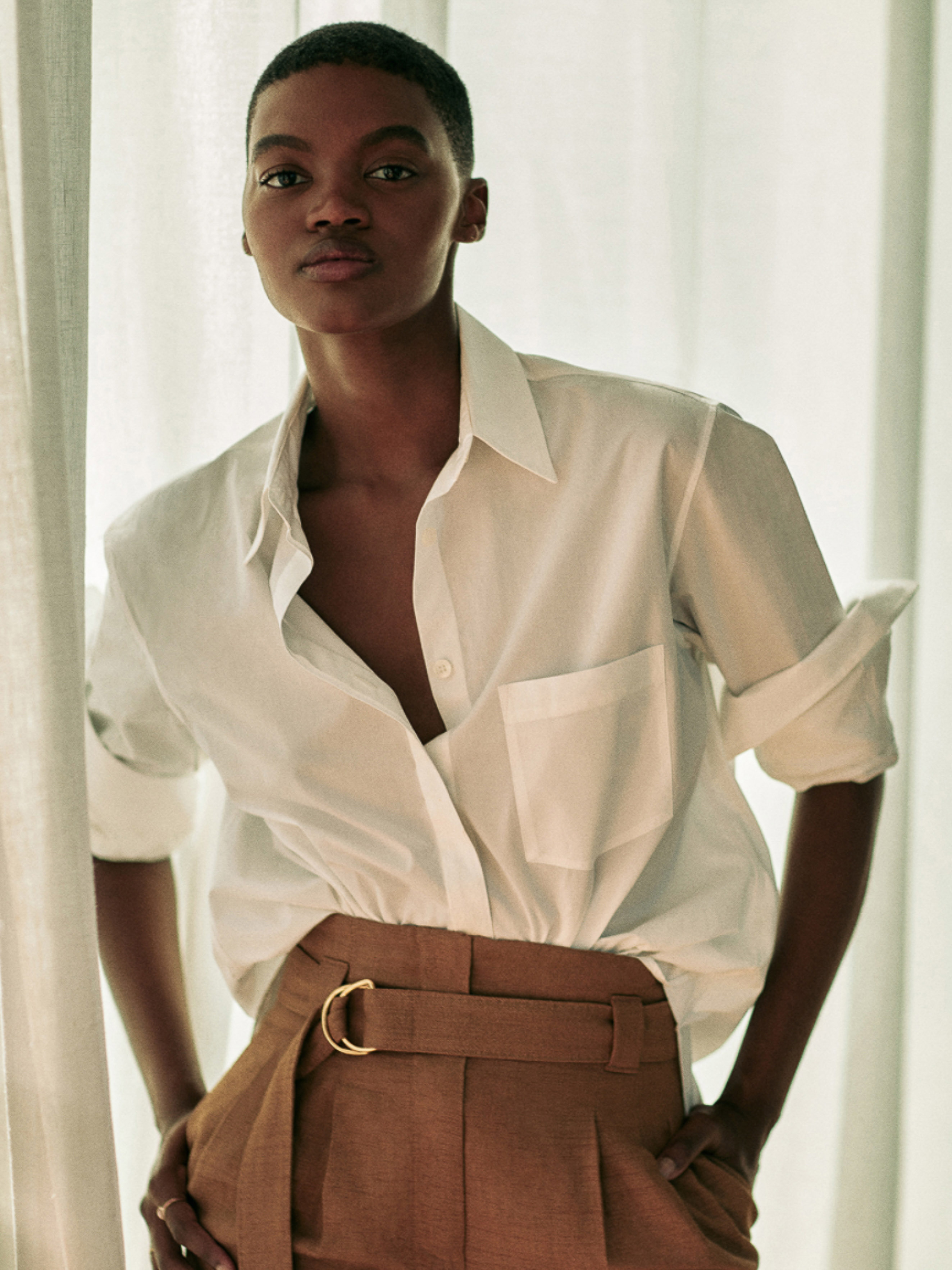 Portrait of a Black female fashion model in a white open button-down shirt and brown trousers, posing indoors in natural light.