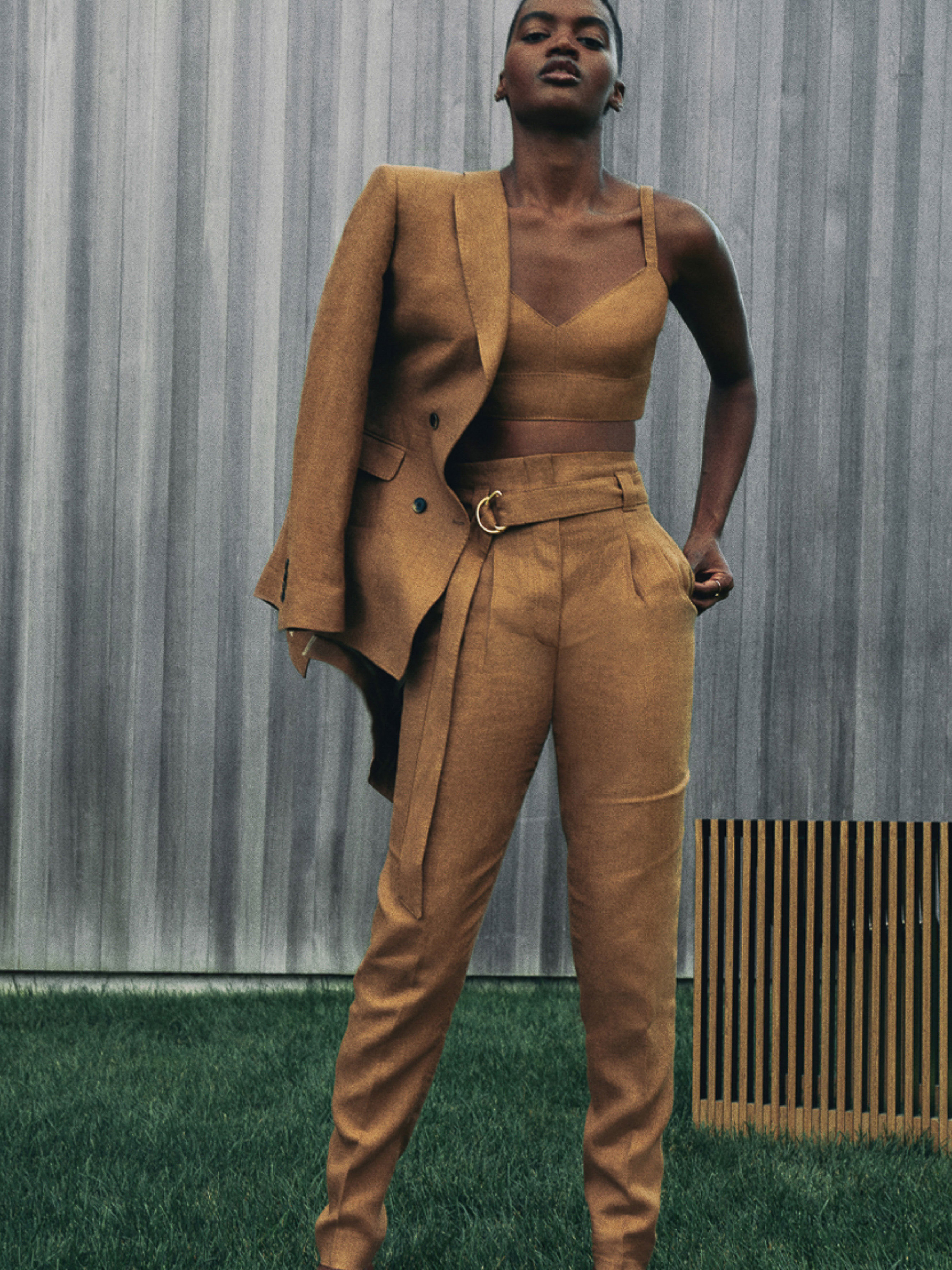 Professional studio shot of a Black woman in a brown linen suit, blazer draped over her shoulder, posing against an outdoor backdrop.