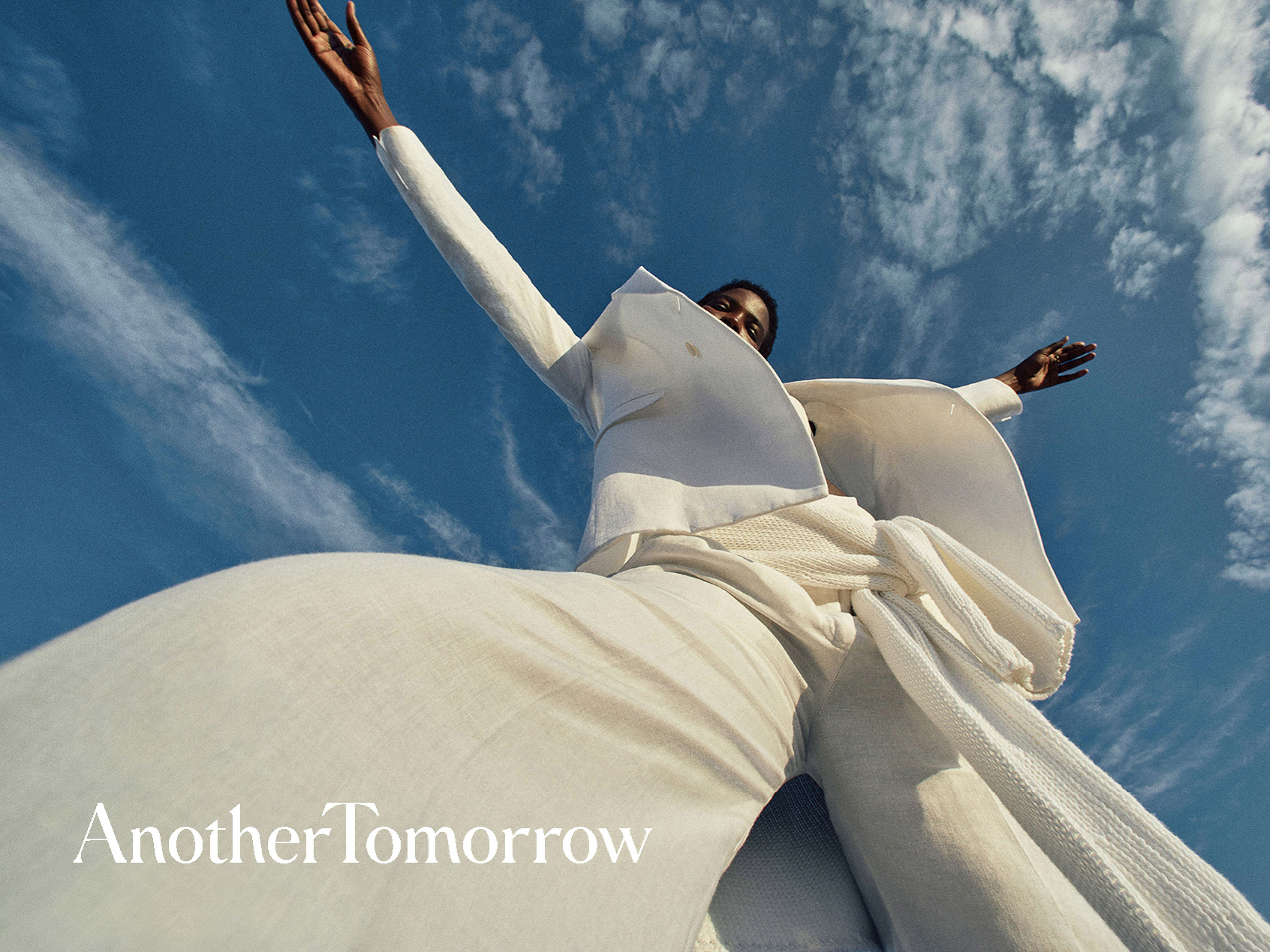 Woman in a white suit stands against a blue sky with wispy clouds, arms outstretched, looking up in a low-angle fashion shot.
