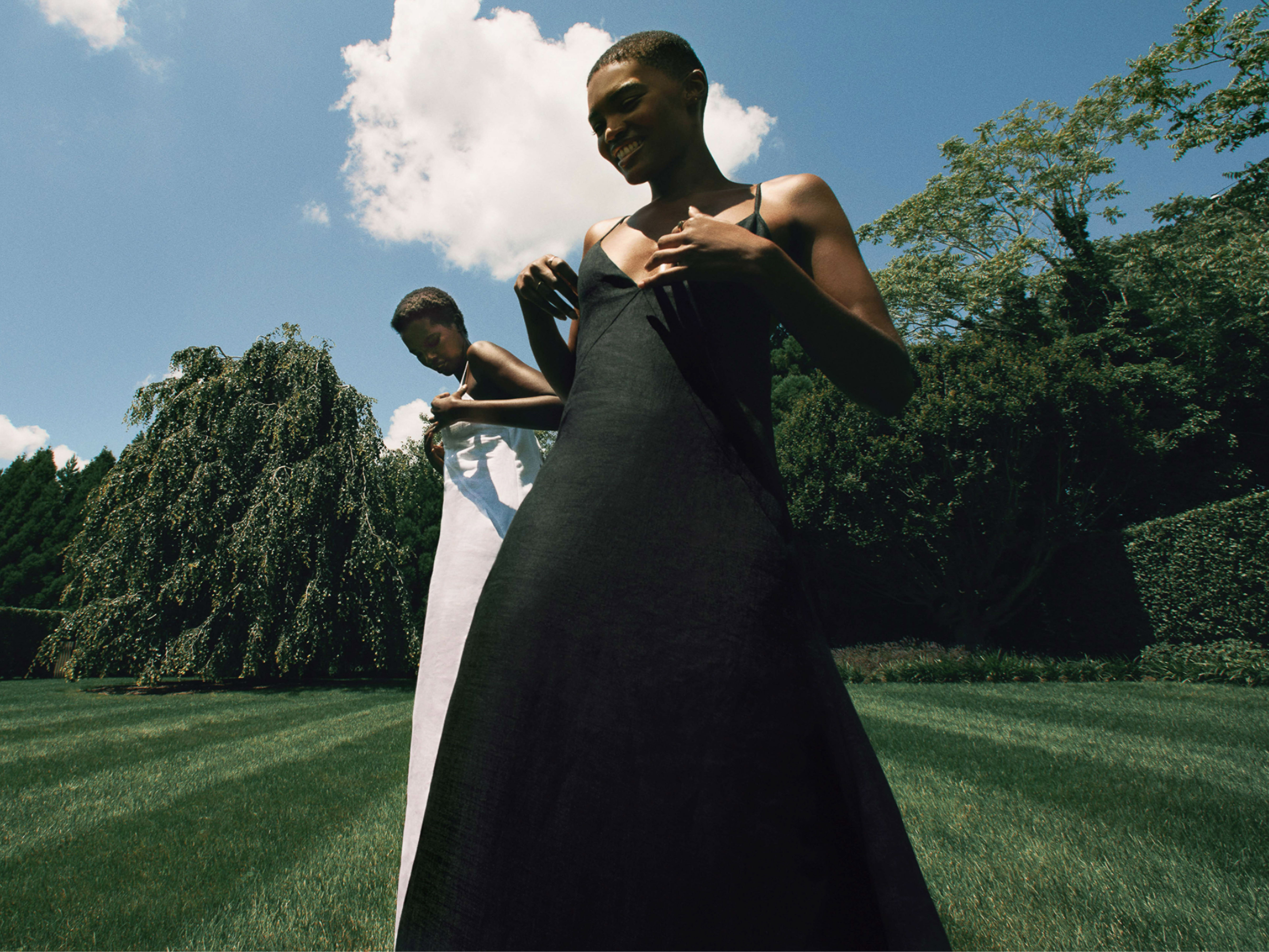 Two Black women posing outdoors on a manicured lawn wearing black and white slip dresses under a sunny blue sky.