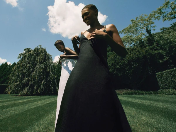 Two Black women posing outdoors on a manicured lawn wearing black and white slip dresses under a sunny blue sky.