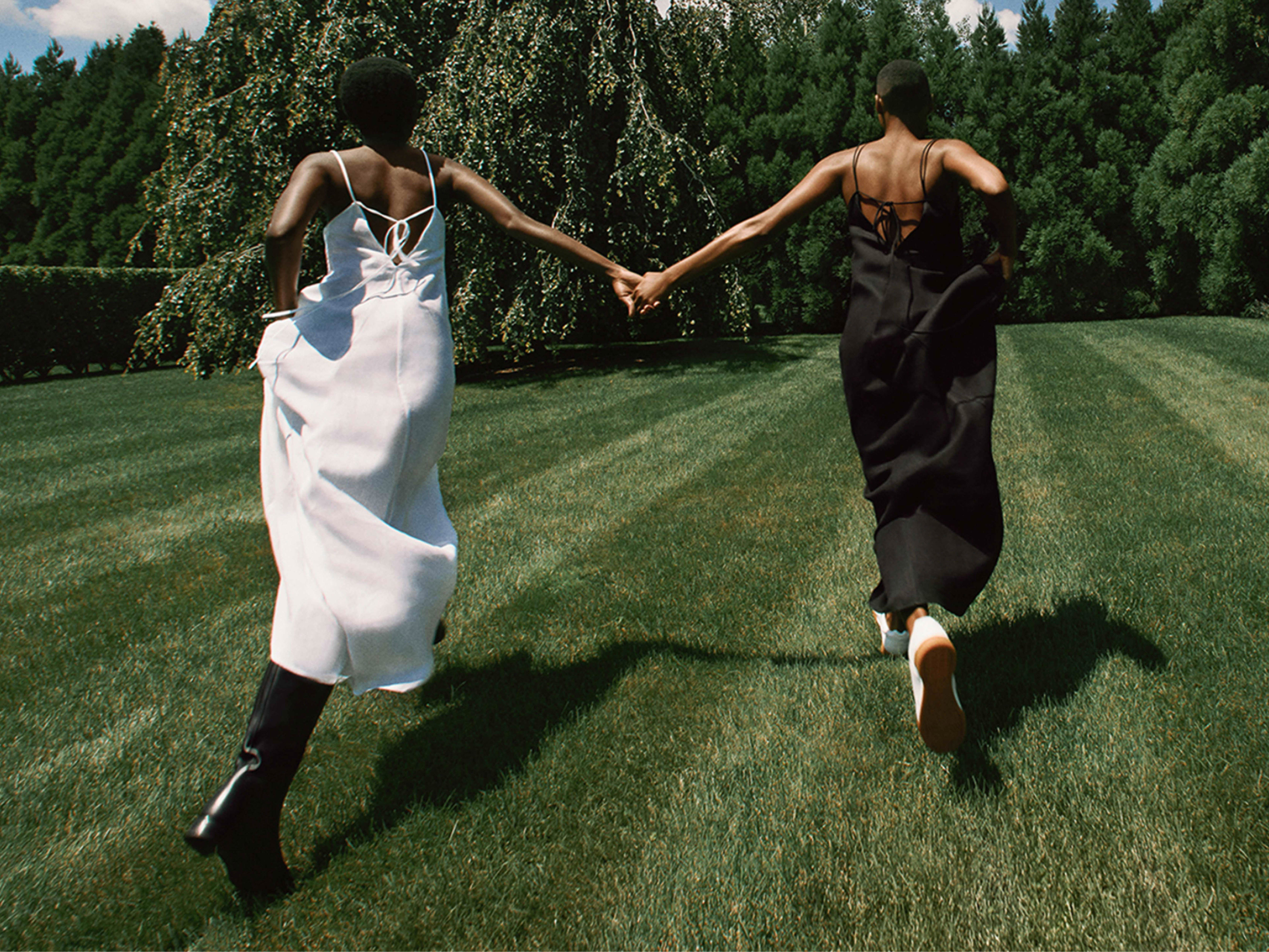 Two adult women in summer dresses run hand-in-hand across a green lawn toward a dense tree line in an outdoor setting.