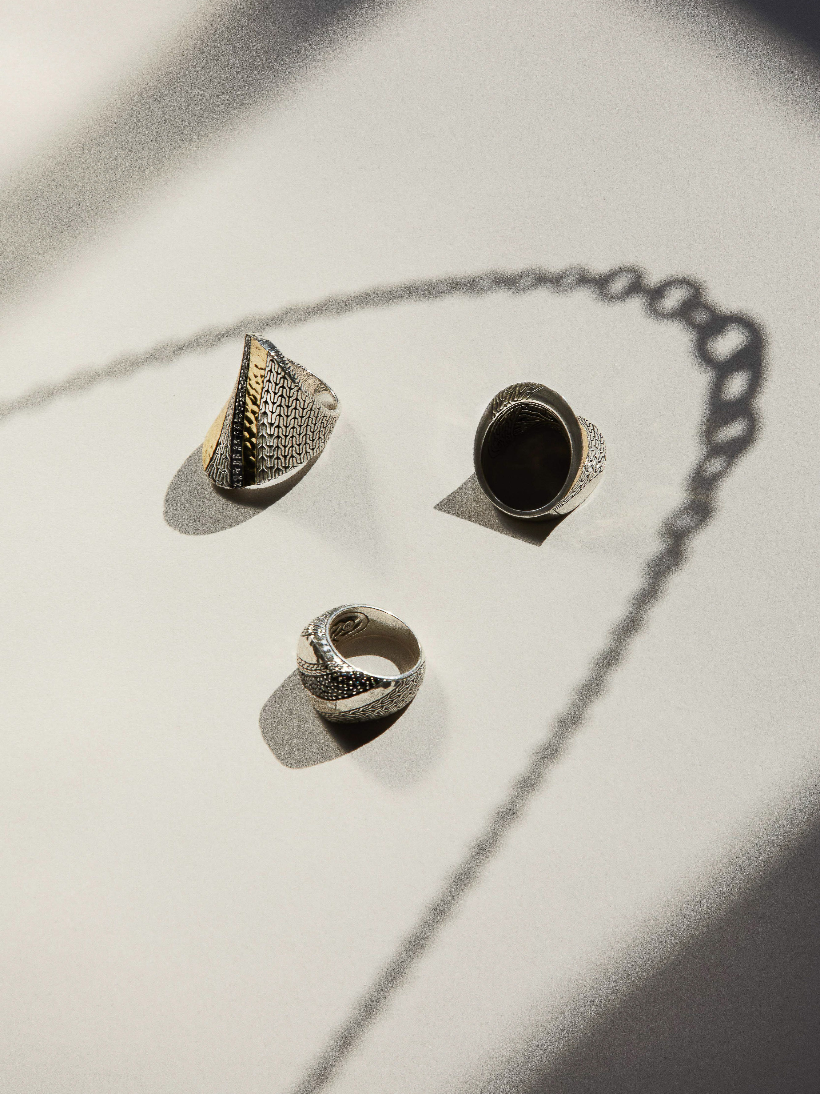 Three silver rings displayed on a white surface with a long shadow cast by a chain-like object, creating a play of light and shadow.