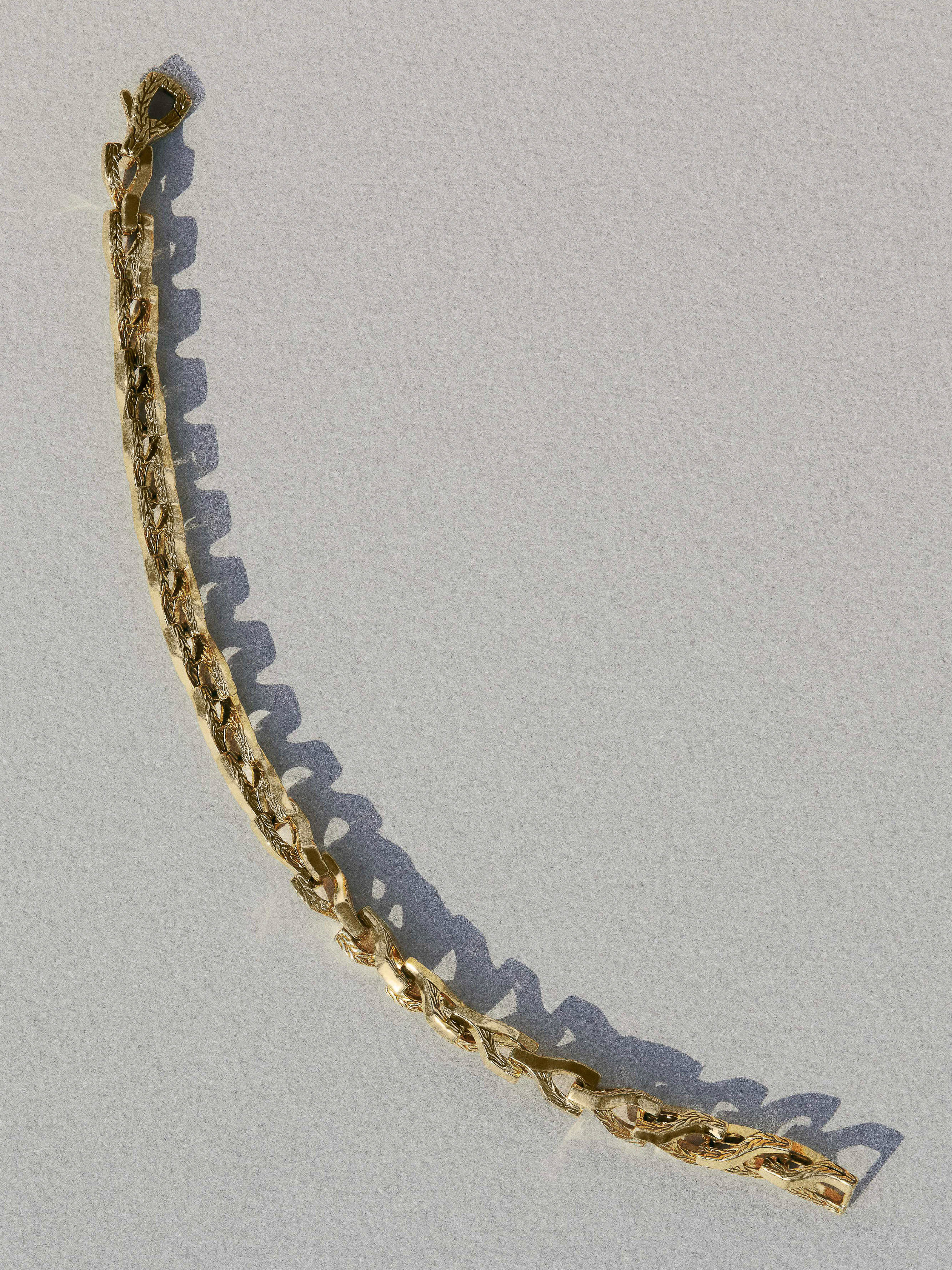 Close-up of an elegant gold metal chain bracelet with intricate weaving design lying flat on a light neutral surface casting soft shadows.