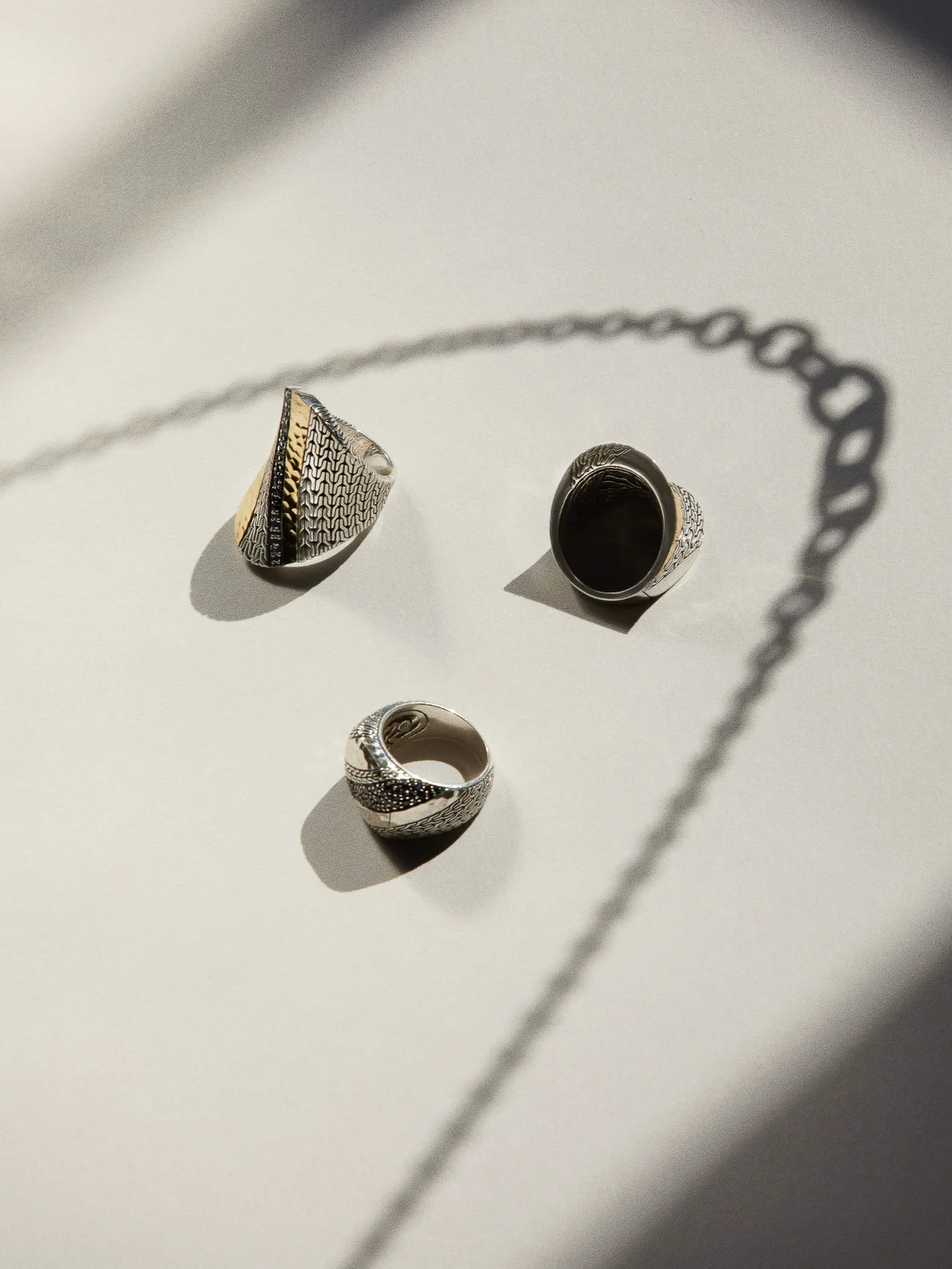 Three textured silver and gold rings arranged on a neutral surface casting sharp shadows beneath a chain silhouette.