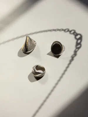 Three textured silver and gold rings arranged on a neutral surface casting sharp shadows beneath a chain silhouette.
