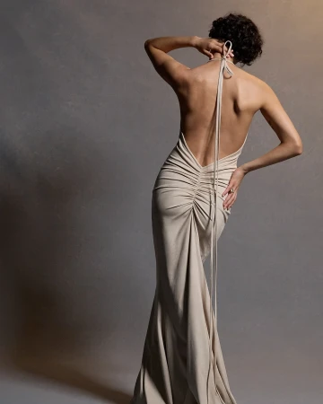 Back view of woman in light brown formal dress with hand on hip in a neutral studio setting with soft lighting and blurred background.