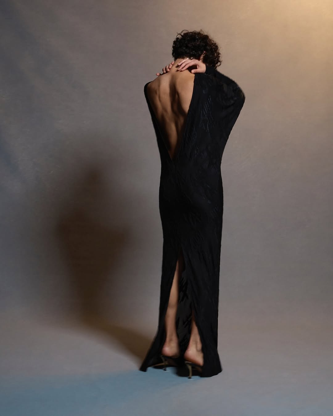 Adult female wearing a long black open-back dress with arms crossed against her shoulders, standing in a neutral-color photo studio.