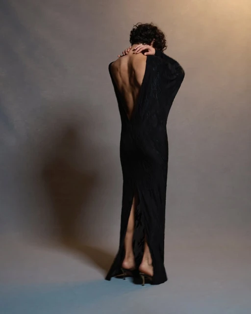 Adult female wearing a long black open-back dress with arms crossed against her shoulders, standing in a neutral-color photo studio.