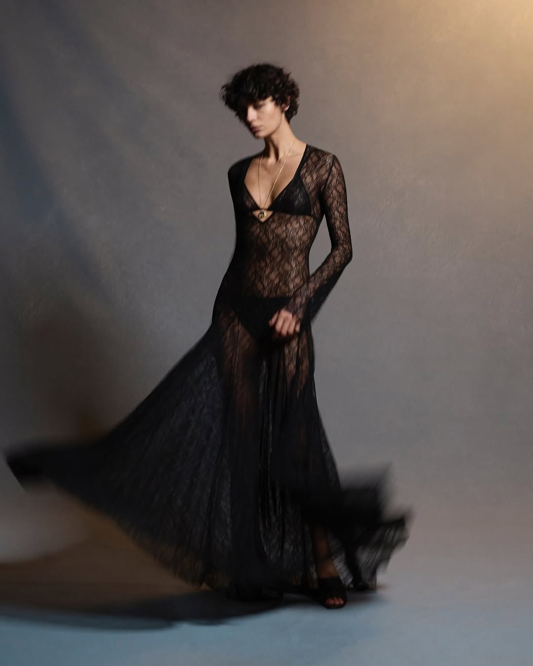 A slender woman poses in a studio wearing a sheer, floor-length black lace dress with a matching bikini underneath against a gray backdrop.