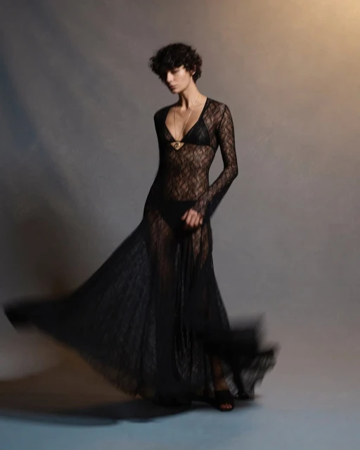 A slender woman poses in a studio wearing a sheer, floor-length black lace dress with a matching bikini underneath against a gray backdrop.