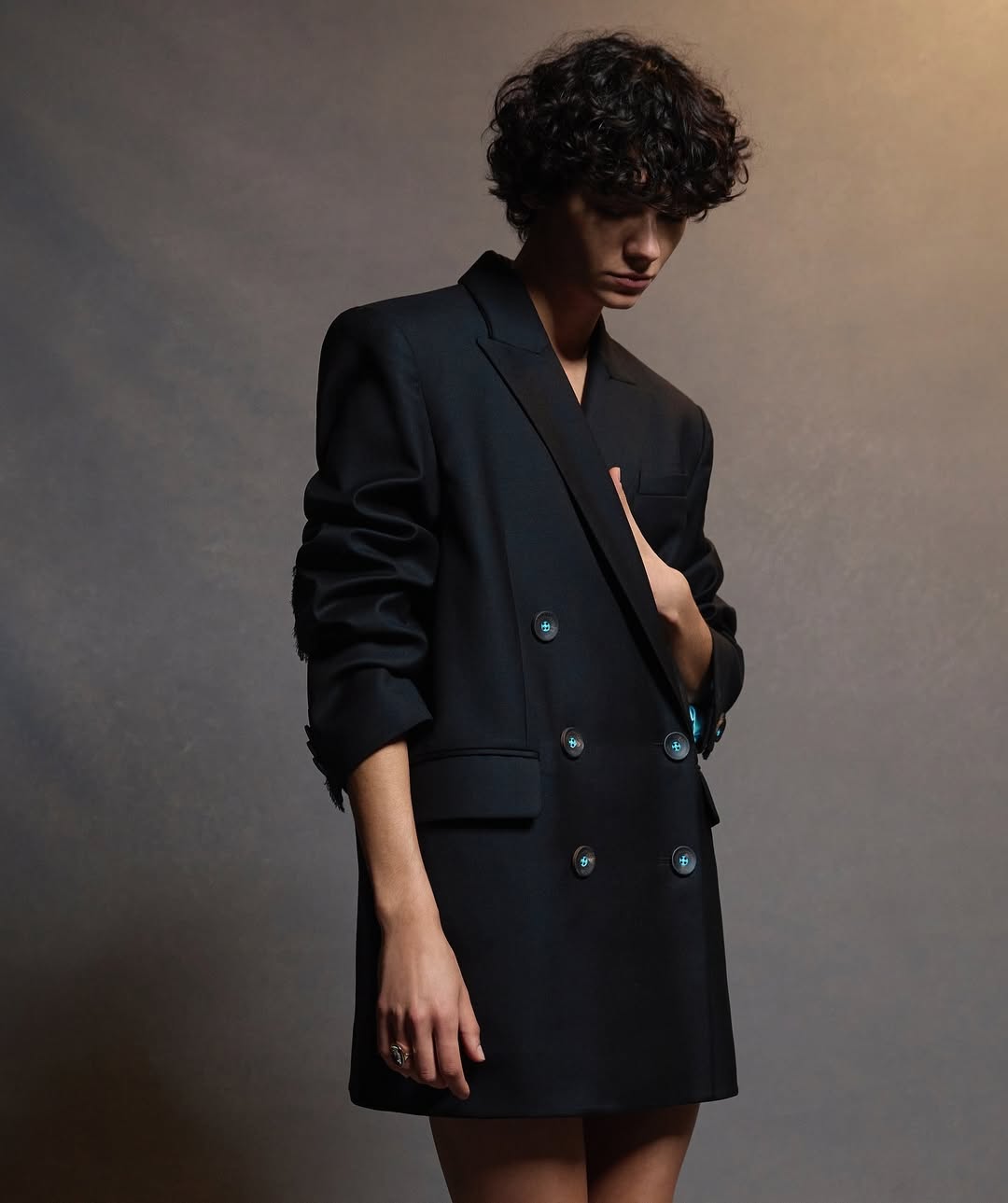 A person with curly hair is wearing a black double-breasted blazer in a studio. The background is a blurry dark neutral color.