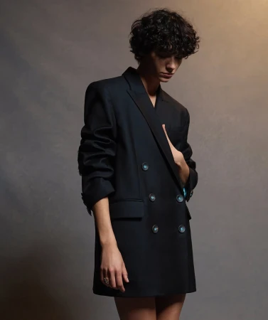 A person with curly hair is wearing a black double-breasted blazer in a studio. The background is a blurry dark neutral color.