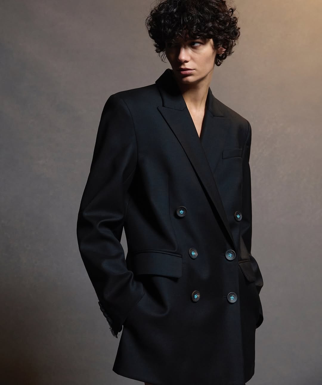 An adult woman with curly hair in a black double-breasted blazer with buttons poses in a studio with neutral lighting, looking at the camera.