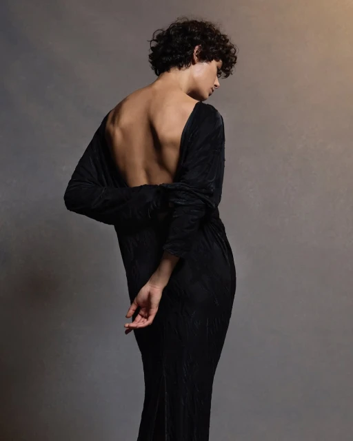 Woman in a long, flowing black dress with an open back, captured in a studio setting with soft, diffused lighting highlighting her form.