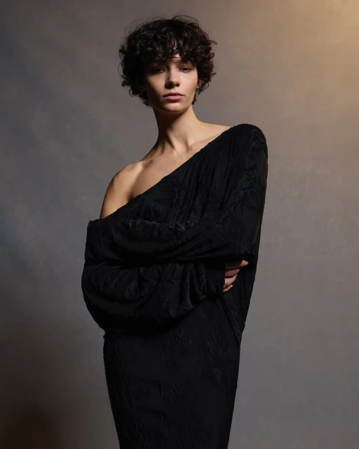 Portrait of a young woman with curly hair wearing a dark off-the-shoulder dress, posing in a studio with a neutral background and soft lighting.