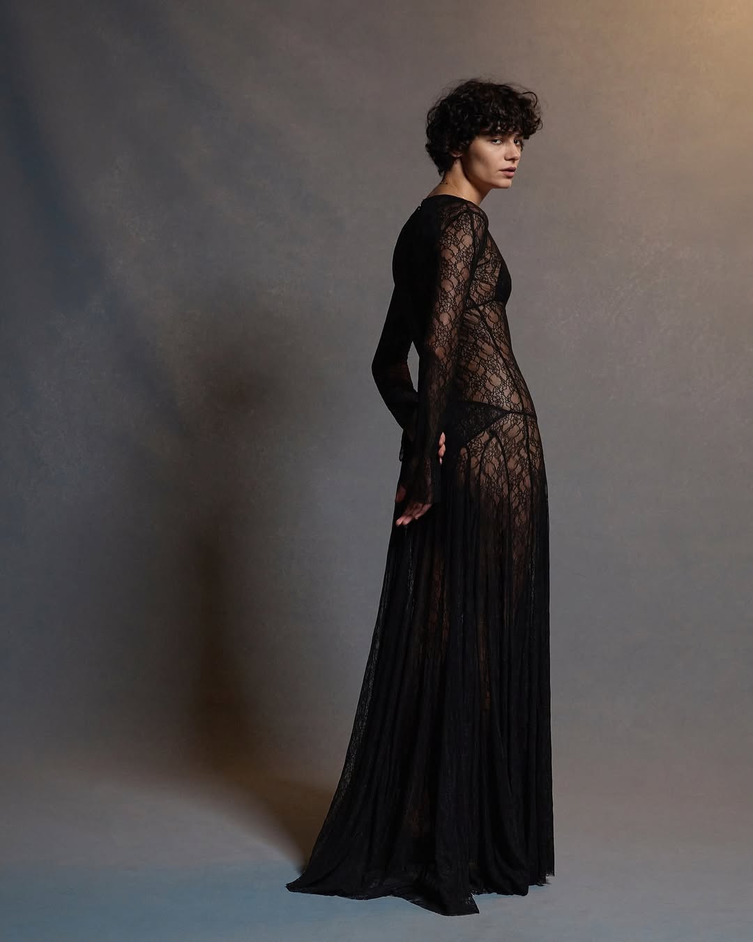A full-length portrait of a woman in a long black lace dress poses against a gray backdrop, illuminated with soft, focused lighting.