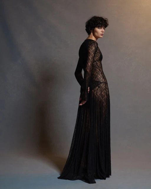 A full-length portrait of a woman in a long black lace dress poses against a gray backdrop, illuminated with soft, focused lighting.