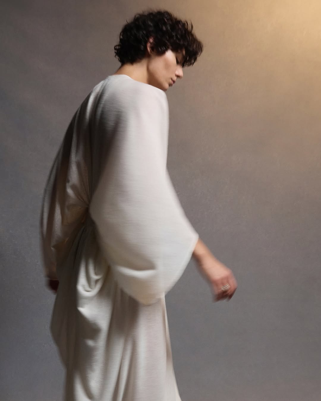 Profile of a curly-haired person in draped white robe-like garment. Soft, neutral background. Fashion, beauty, and style are highlighted.