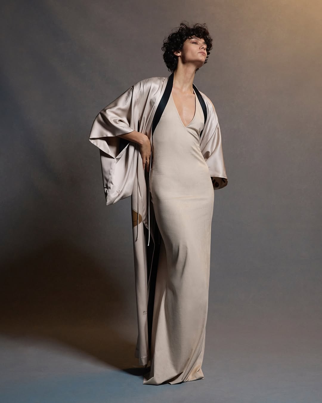 A woman in a full-length dress and robe poses in a studio, looking off-camera with soft lighting and a neutral color palette.