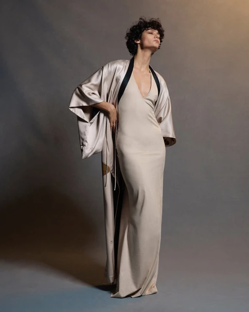 A woman in a full-length dress and robe poses in a studio, looking off-camera with soft lighting and a neutral color palette.
