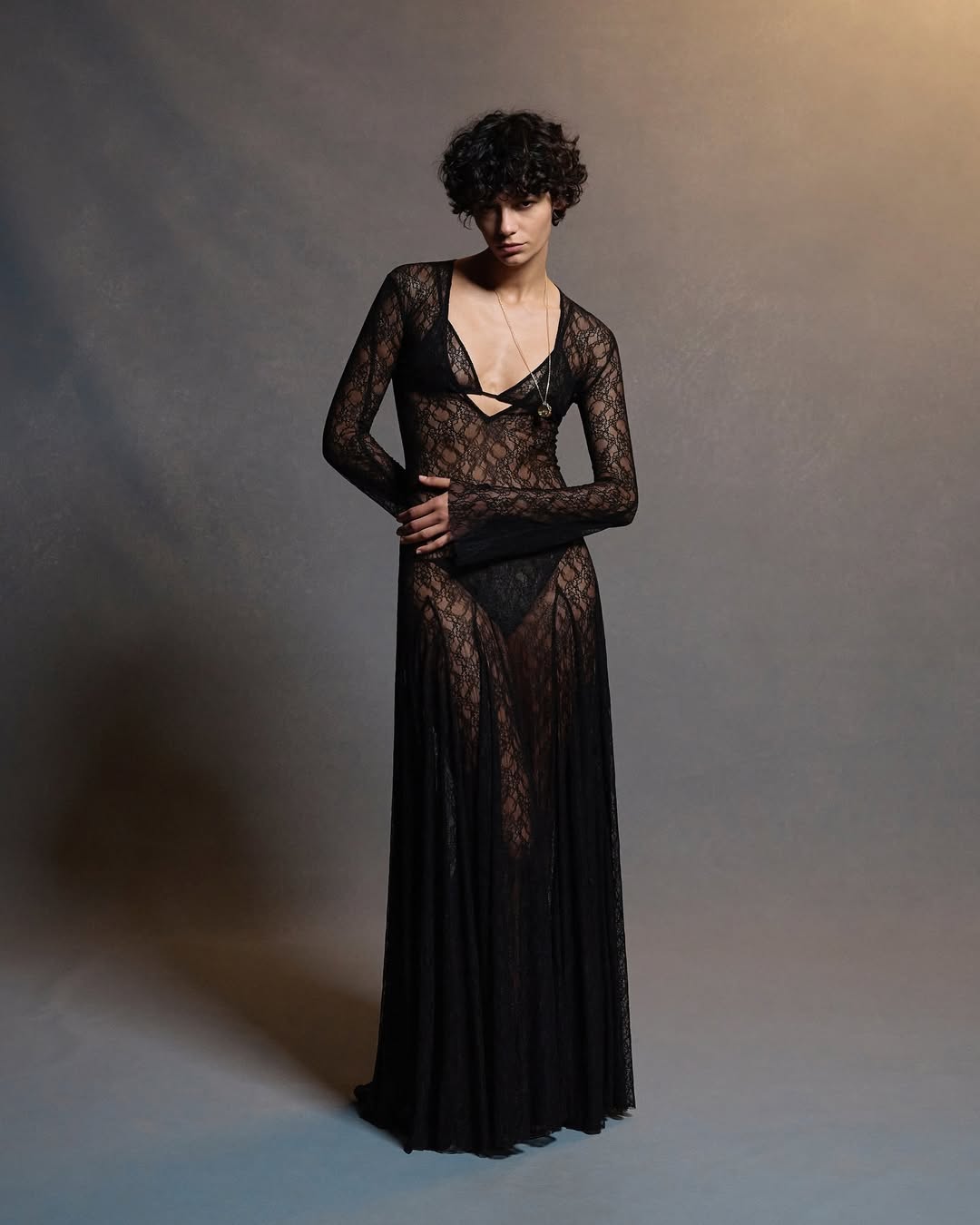 Full-length shot of a model with short curly hair wearing a long black lace dress in a studio with a soft, focused light.