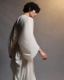 A man with dark curly hair wearing a white flowing robe walks against a mottled grey studio backdrop with warm lighting.