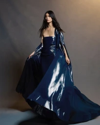 Model wearing a glossy dark blue strapless gown and matching cape against a textured grey studio backdrop.